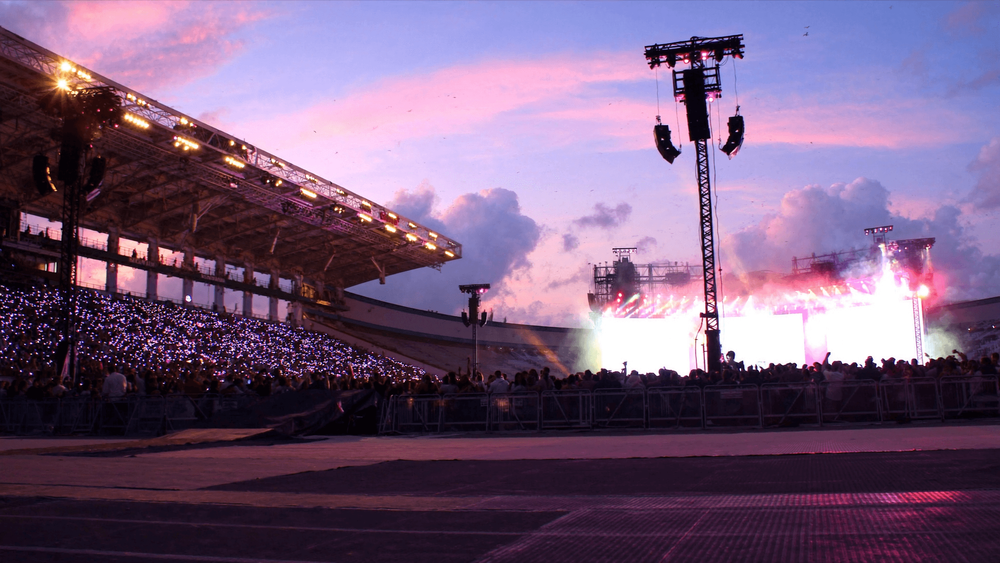 Open-air stadium concert at dusk, with a glowing stage and thousands of phone lights in the stands under a pink sky