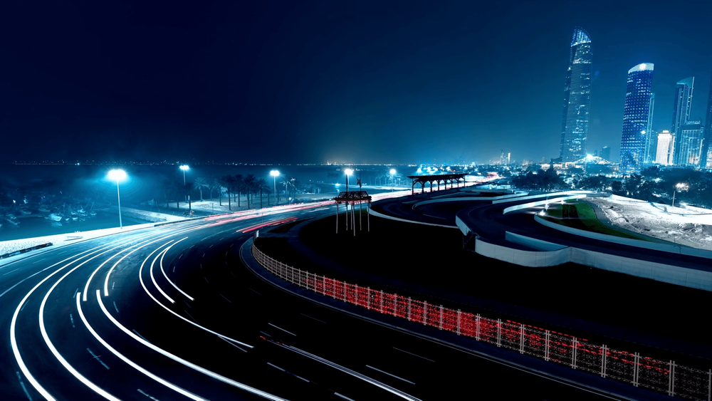 Night street circuit beside a marina, with light trails sweeping through a floodlit curve below a glowing city skyline