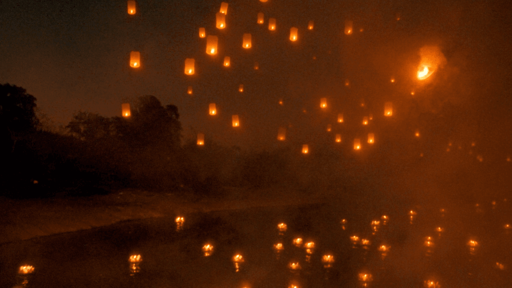 Glowing paper lanterns rising into the night sky over a river during the Yi Peng festival in Chiang Mai