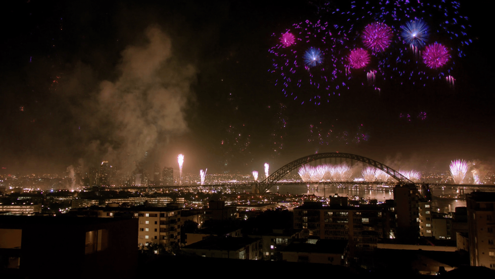 New Year's Eve fireworks bursting over a harbour city and an arch bridge at midnight