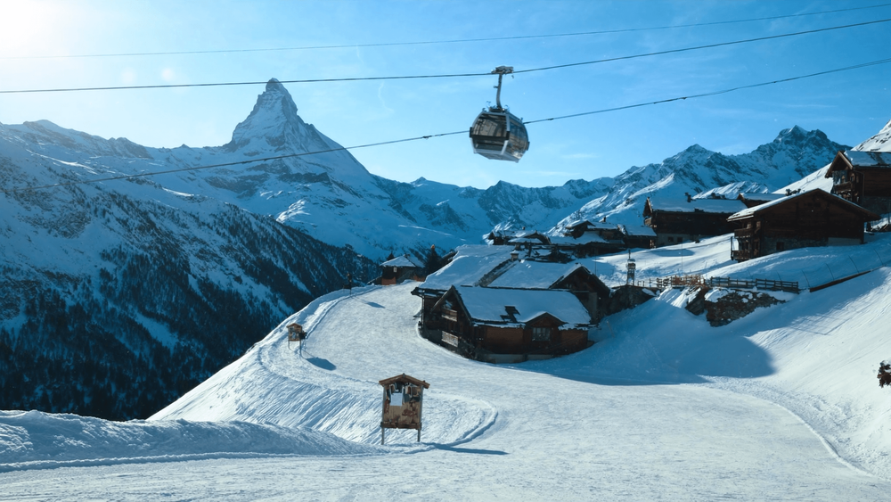 Alpine ski resort on a sunny morning, with a gondola above a groomed piste, wooden chalets and a sharp snow-covered peak