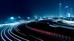Night street circuit beside a marina, with light trails sweeping through a floodlit curve below a glowing city skyline